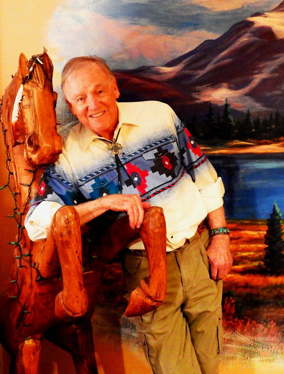 Ed Breeding leaning on a carved wooden horse in front of one of his mountain landscapes